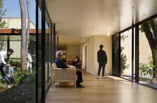 An elderly man sits in a chair looking outside as a woman stands nearby inside hospice.