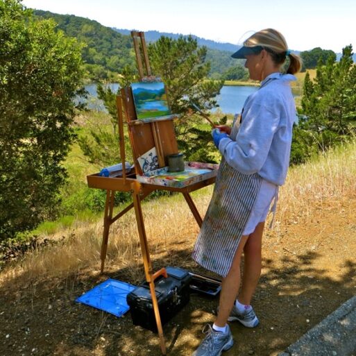 Author Dana Shields painting at an easel near a lake.