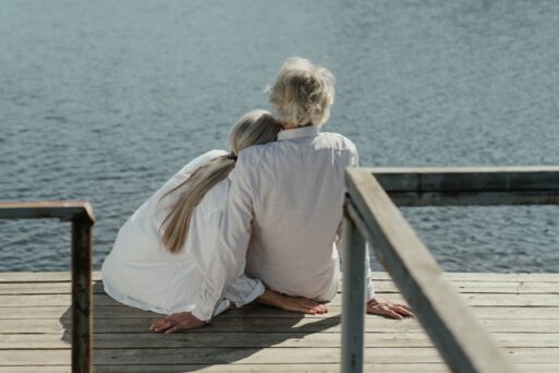 An elderly couple embrace on a dock while gazing out over the water.