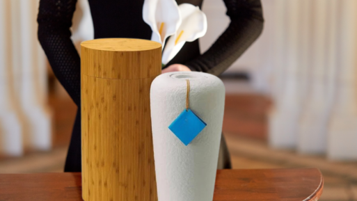 Two urns - one made of bamboo and the other some sort of gray, biodegradable material, are sitting on a table in the foreground. Behind them is a woman's torso, dressed in black and holding white lillies.