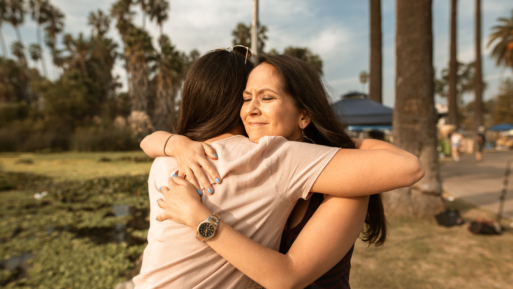 Two women embrace tearfully, with tropical trees in the background.