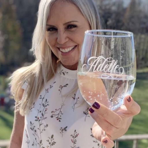 A blonde woman smiles as she holds up a stemless wine glass with the name Adele etched into it