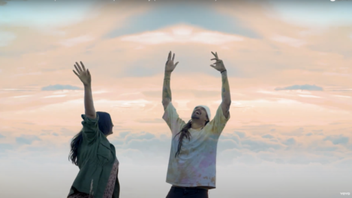 A man and a woman in casual clothing are dancing with their hands in the air in front of a pastel sky