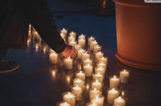 A collection of votive candles are lit and assembled in loose rows in a dark room