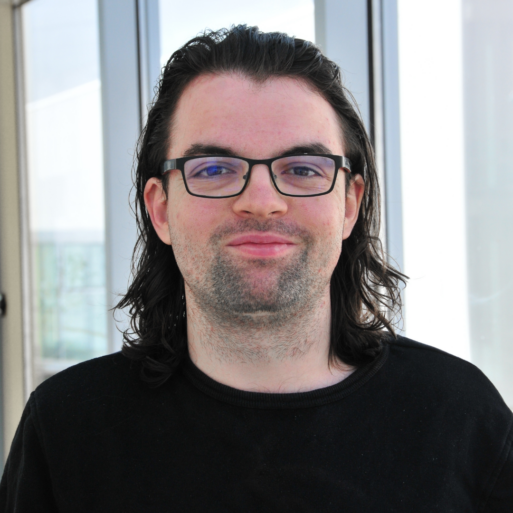 A man with long dark hair and glasses smiles proudly for the camera in front of bright office windows