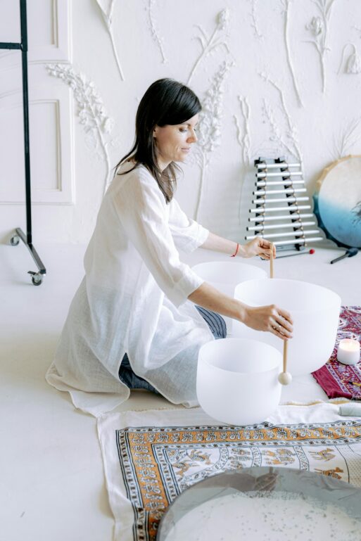 A woman in an all-white room (and dressed in loose, flowing, white clothing) plays live music on a set of crystal bowls with wooden mallets