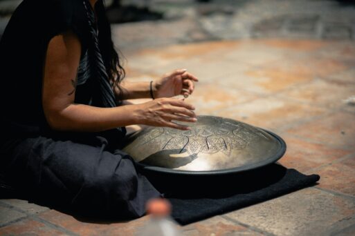 A woman with long, wavy hair sits cross-legged on the floor as she plays the hang drum, a metal, oblong-shaped instrument