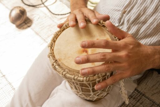 A man dressed in light, neutral-toned clothing holds a hand drum to his lap as he plays it with his fingers