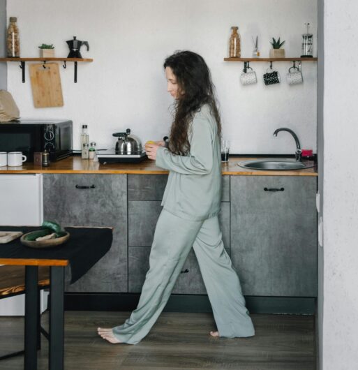 A woman walks a lonely kitchen in her pajamas.