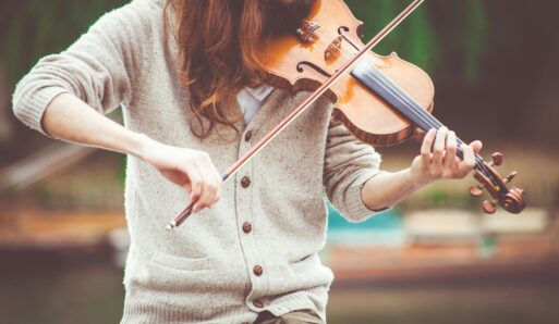 A woman with long, brown hair is playing the violin while wearing a neutral-tone, button-down cardigan