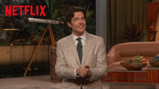 Comedian John Mulaney - a caucasian white man with floppy brunette hair and a handsome face - delivers his opening monologue for his show on a set decorated like a livingroom.