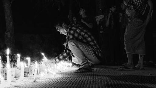 A man in a plaid shirt crouches down to light a candle at a vigil
