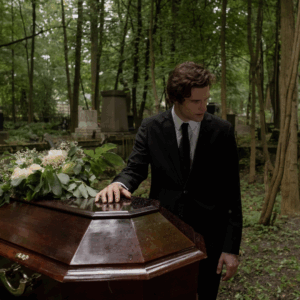 A man in a black suit places his hand on a casket and looks away. They are in a wooded graveyard, and there are fresh flowers on the casket.