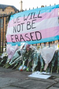 Flowers leaned against a fence under a trans flag with "We will not be erased" written across it