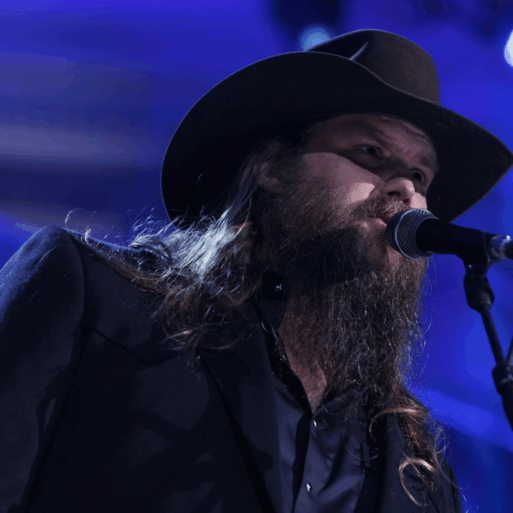 Chris Stapleton (a white man wearing a black cowboy hat and has a long, dark brown beard) is singing into a microphone on stage