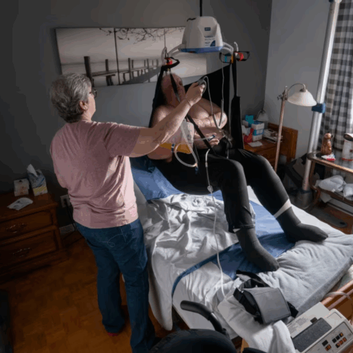 A woman with short, gray hair assists a man in a medical sling over a hospital bed