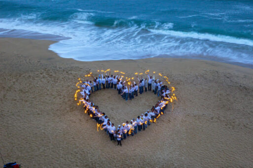 In an aerial photograph, people are formed in a heart shape on a beach and hold sparklers in their hands.