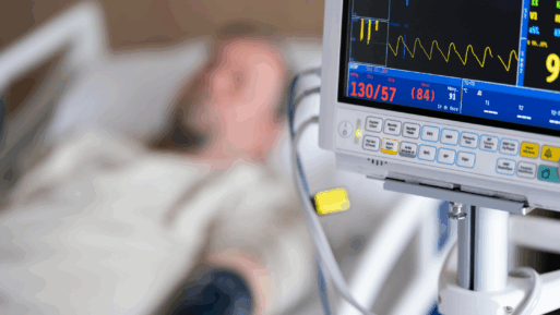 A man is lying in a hospital bed, but blurred because focus is on a medical monitor that is in the foreground