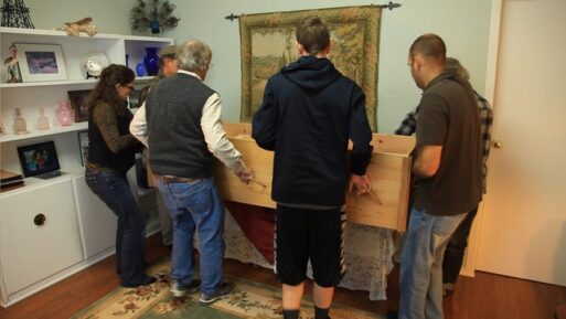 A family surrounds a wooden casket in a room at home, preparing to lift it.