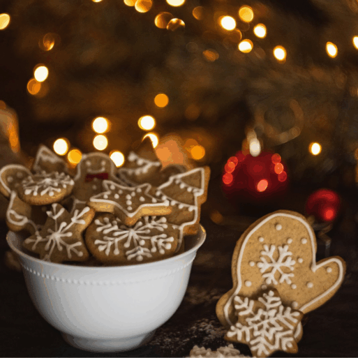 A bowl of beautifully decorated gingerbread cookies is in front of a christmas tree. The cookies are all shaped like mittens, trees, or snowflakes, and decorated with white icing.