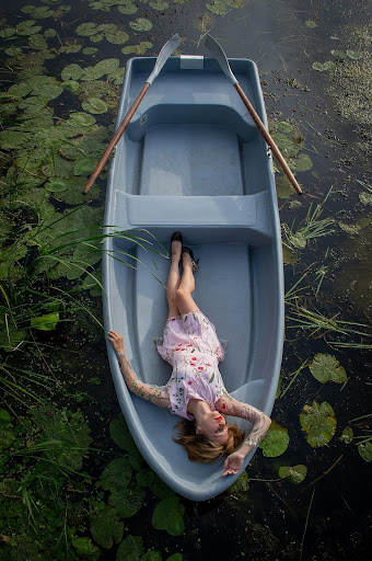 person in dress and heels reclining in a row boat surrounded by lily pads