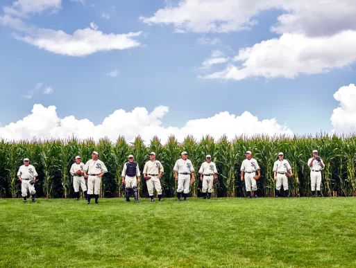 A row of men dressed in old-fashioned White Sox baseball uniforms stand in front of a verdant field of corn stalks