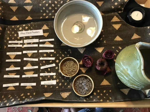 On a small table, sacred items are laid out as an altar for the dead. There is a bowl of water in a ceramic dish, dried roses, dried herbs like lavender and rosemary in little bowls, and a candle. There are scraps of paper with the names of people who have been killed, under a label that says, "#saytheirnames."