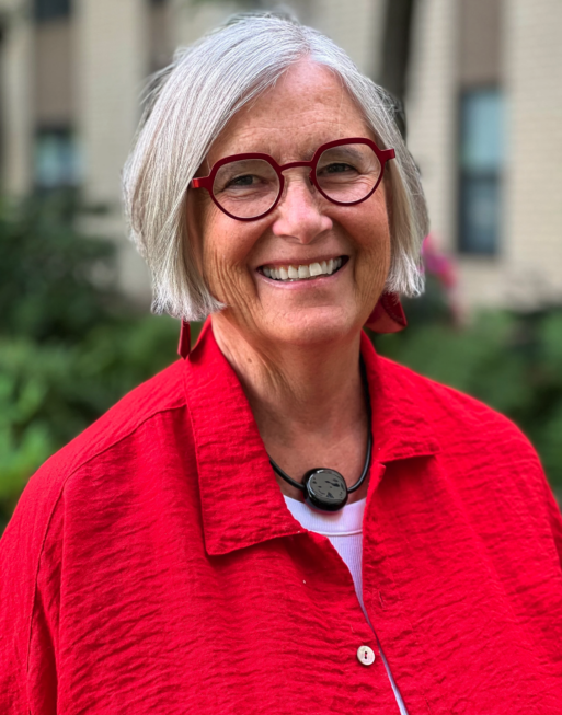 A woman with grey hair cut in a blunt bob is smiling for the headshot photo, wearing a vibrant red shirt, funky glasses and a chunky necklace. 