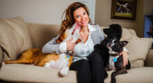 Dr. Dani McVety sits on a couch surrounded by three dogs. One is licking her chin while the other two gaze up at her.