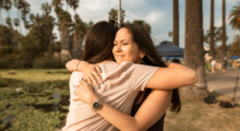 Two women embrace tearfully, with tropical trees in the background.