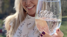 A blonde woman smiles as she holds up a stemless wine glass with the name Adele etched into it