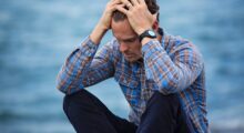 Man holding head in emotional distress by a body of water.