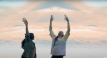 A man and a woman in casual clothing are dancing with their hands in the air in front of a pastel sky