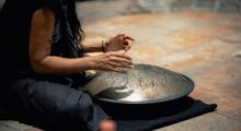 A woman with long, wavy hair sits cross-legged on the floor as she plays the hang drum, a metal, oblong-shaped instrument